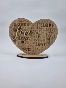 Freestanding wooden heart card engraved with an all-over “LOVE” pattern and raised script reading “I Love Us,” displayed on a stand.