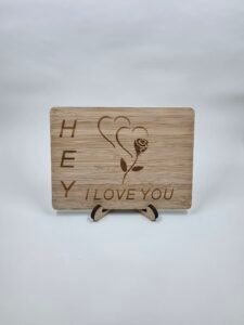 Wooden postcard keepsake engraved “Hey I LOVE YOU” with rose and heart design, displayed on a small stand against a red background.