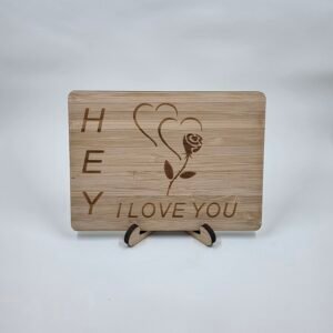 Wooden postcard keepsake engraved “Hey I LOVE YOU” with rose and heart design, displayed on a small stand against a red background.