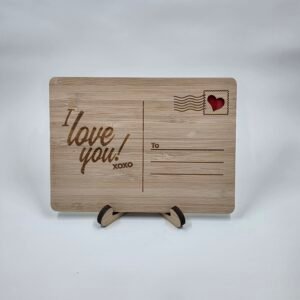 Wooden postcard keepsake engraved “I Love You! xoxo” with stamp detail and lines to write loved one's name, displayed on a small stand.